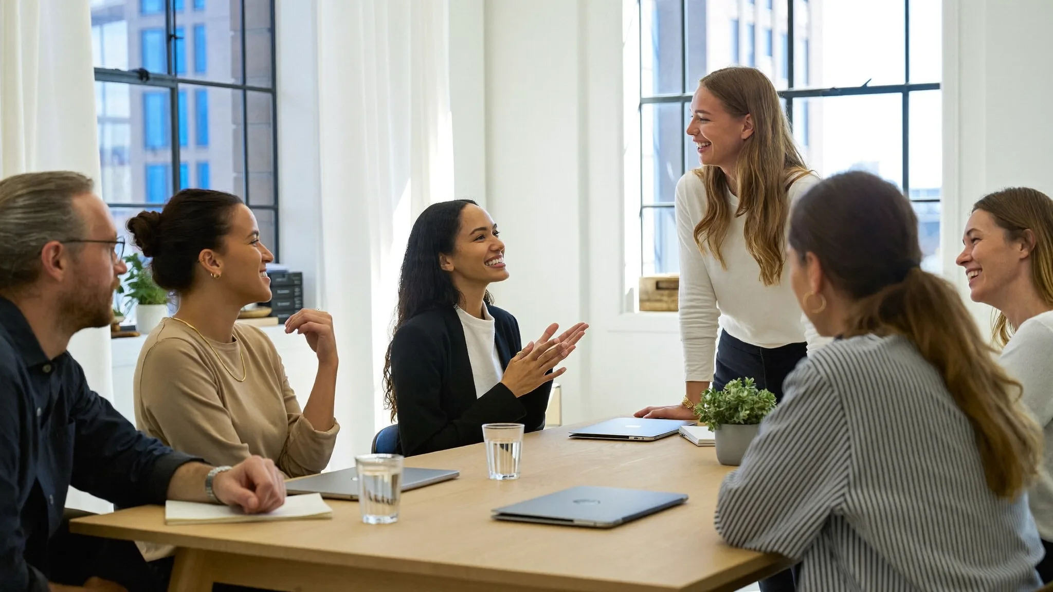 Six entrepreneurs around a table in a bright, daylit studio, one standing mid-sentence while the others listen and laugh